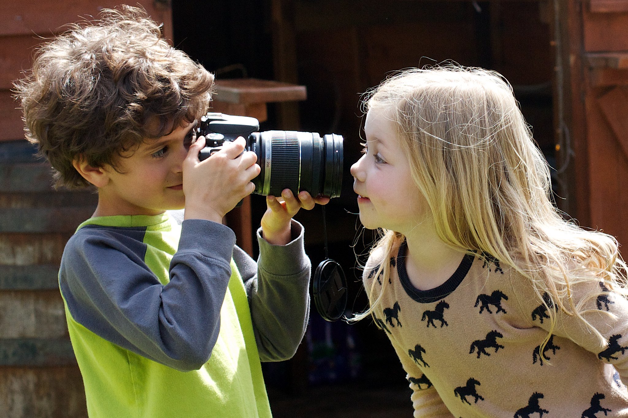 A boy looks through a large camera at a smiling girl right in front of the lends.