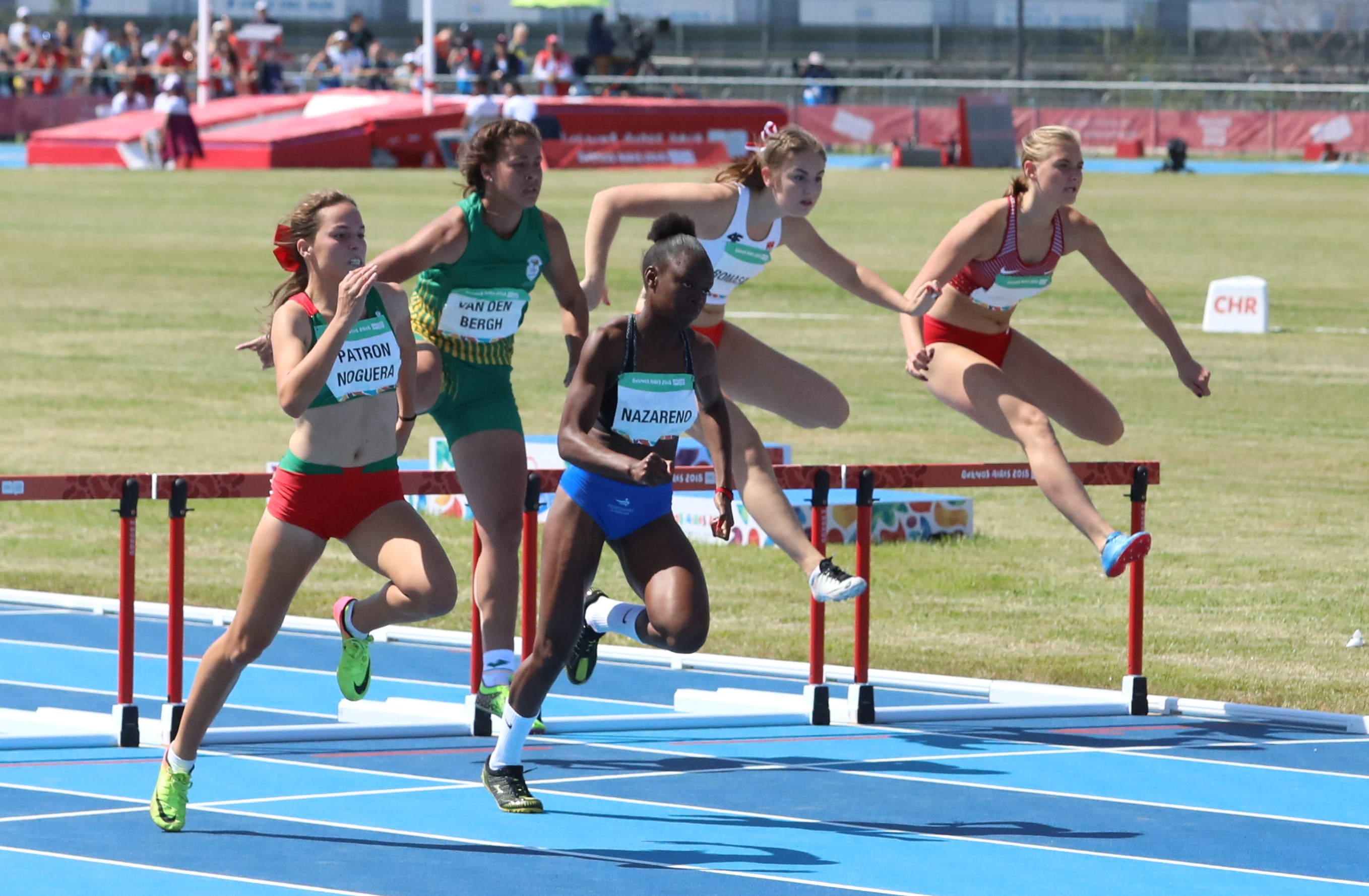 Photo of five runners jumping over hurdles.