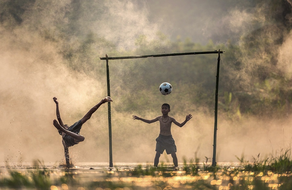 Photo of children playing soccer in a wet field with one boy falling after kicking the ball and another boy trying to block the ball with his head. 