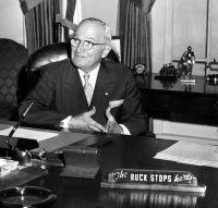 Harry Truman poses for a photograph at the recreation of the Truman Oval Office at the Truman Library in 1959.  On the wodden desk is the famous sign "The Buck Stops Here".