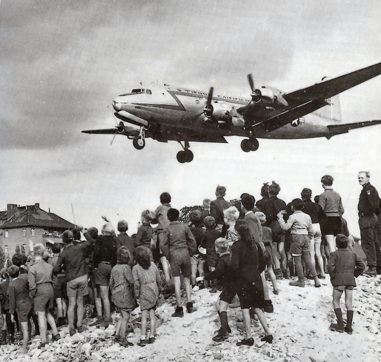 A crowd of people, mostly children, stand on a hill.  Their backs are to the camera as they look up towards an airplane that is coming down for landing close by.