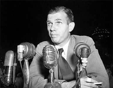 Alger Hiss wears a suit and tie, holds a cigarette and prepares to speak from behind a row of microphones.