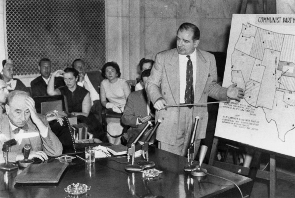 McCarthy stands with a pointer stick in front of a map of the U.S. A partial heading of the map can be seen, reading "Communist Party." Several people in the courtroom are holding their heads and looking away from McCarthy, including Joseph Welch, whom McCarthy is facing.