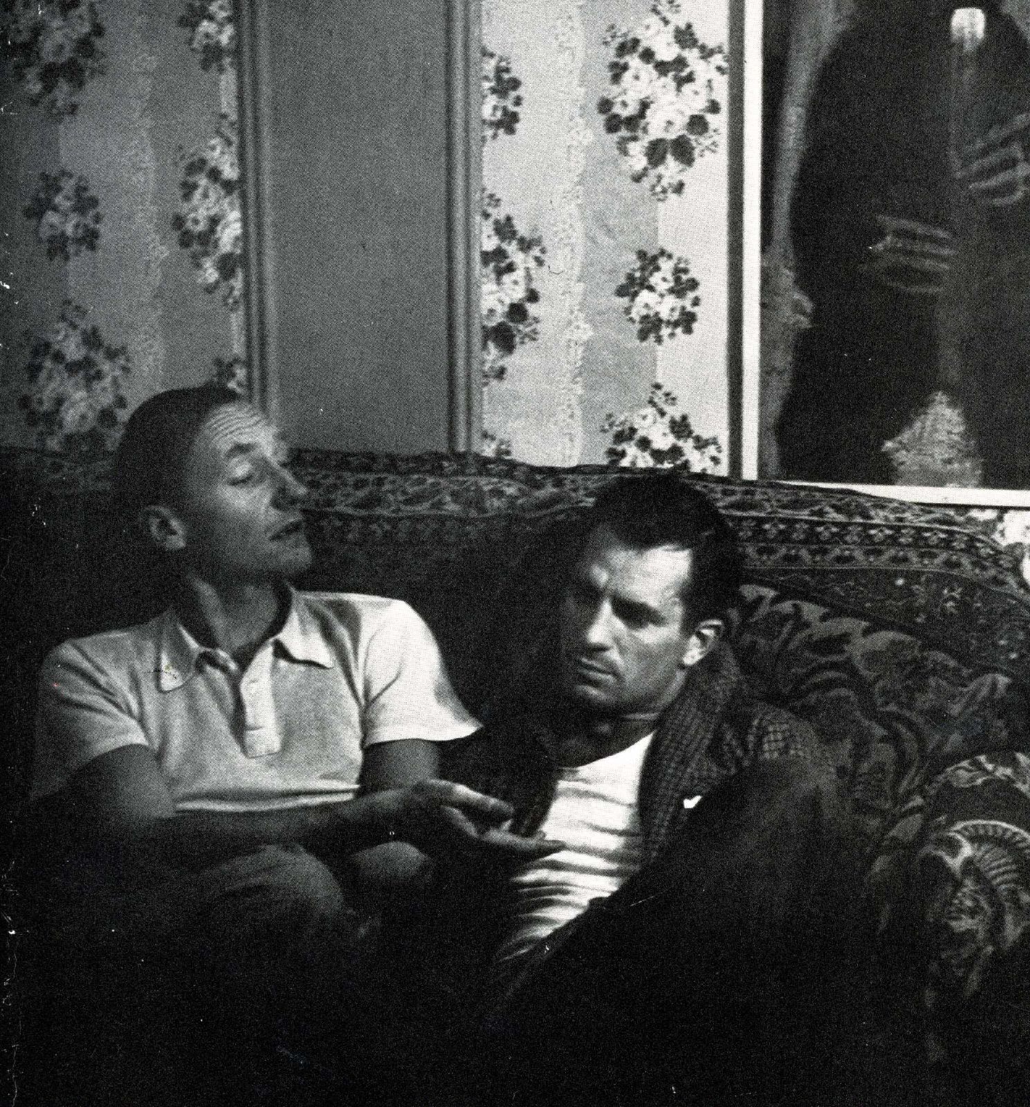 Two men sit on an oriental patterned couch. Ginsberg is on the left, turning towards Kerouac and raising his eyebrows.