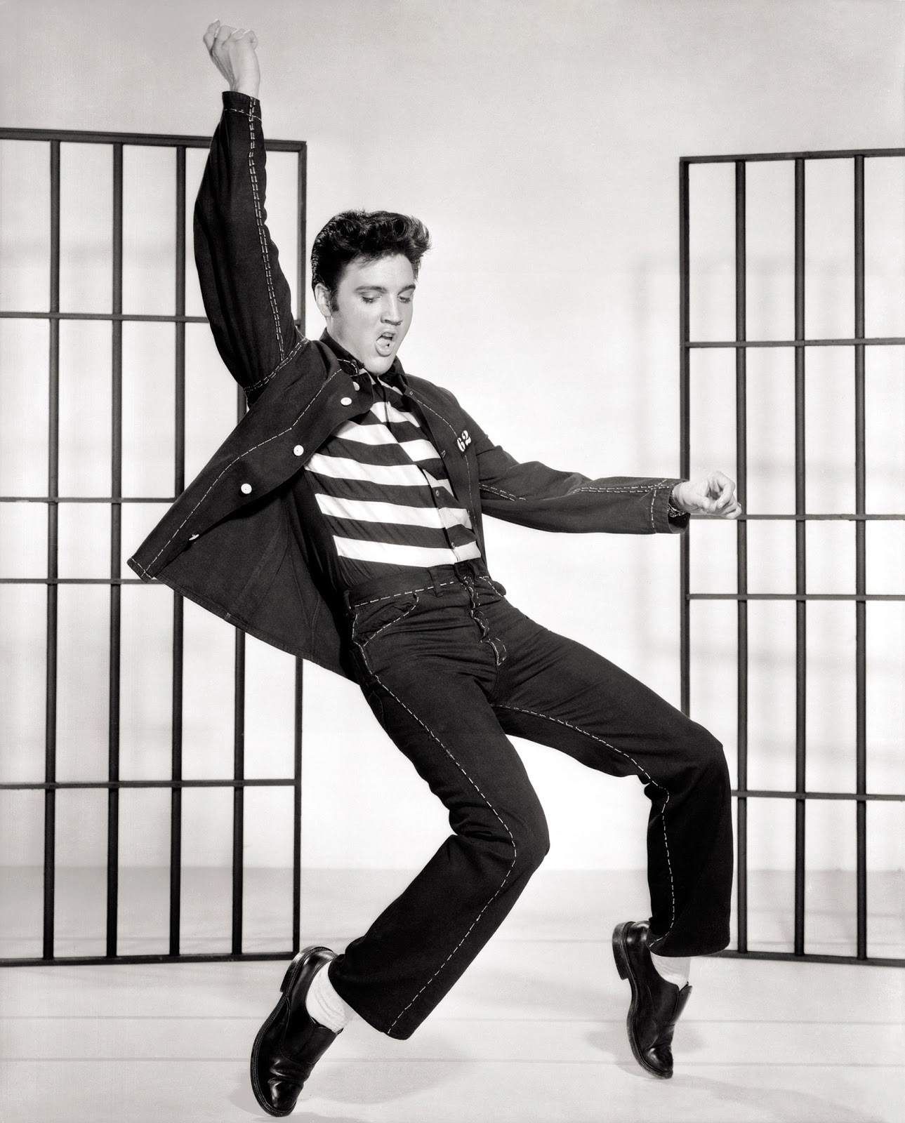 Elvis Presley in a striped T-shirt and matching jeans/jacket set, executes a dance move on tip-toe in front of movie-set jail bars.