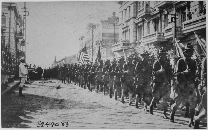 American troops in Vladivostok parading before the building occupied by the staff of the Czecho-Slovaks. Japanese marines are standing at attention as they march by. Siberia, August 1918. By Underwood & Underwood (U.S. National Archives and Records Administration) [Public domain], via Wikimedia Commons