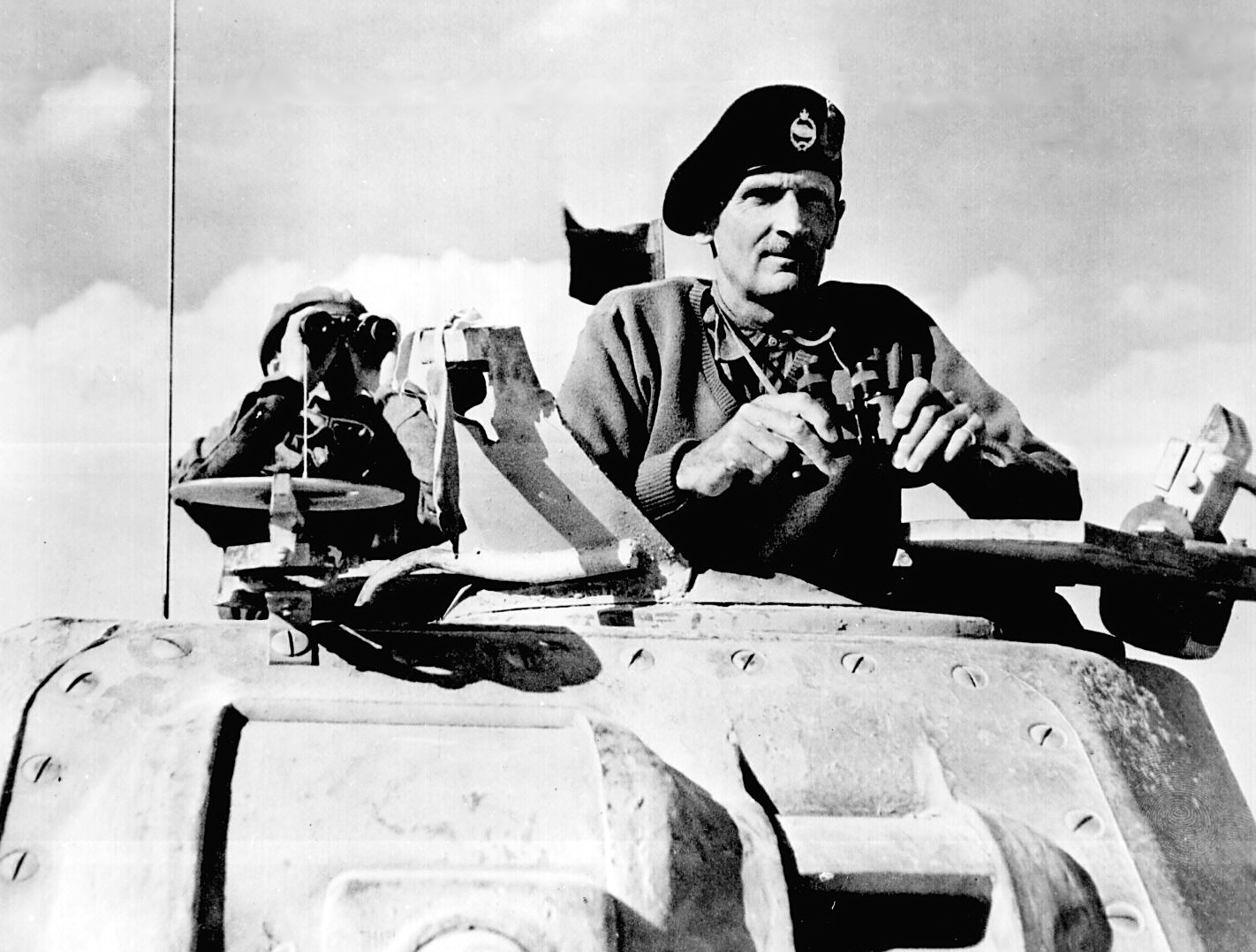 General Montgomery peers out from the front hatch of an army tank, holding binoculars.  Another person is also watching from the tank in a hatch behind Montgomery; this person is holding binoculars up to his eyes.