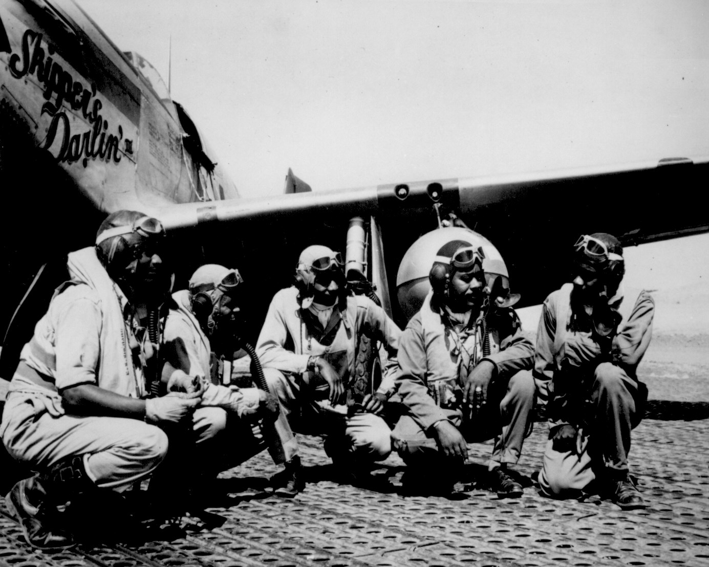 5 Tuskegee Airmen in pilot's gear crouch in front of a fighter plane.