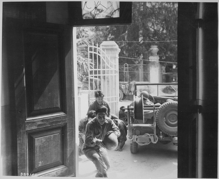 Photo shot from inside a building as soldiers rush in through the doorway for cover. An army vehicle is visible outside, as well as an arched metal gate.