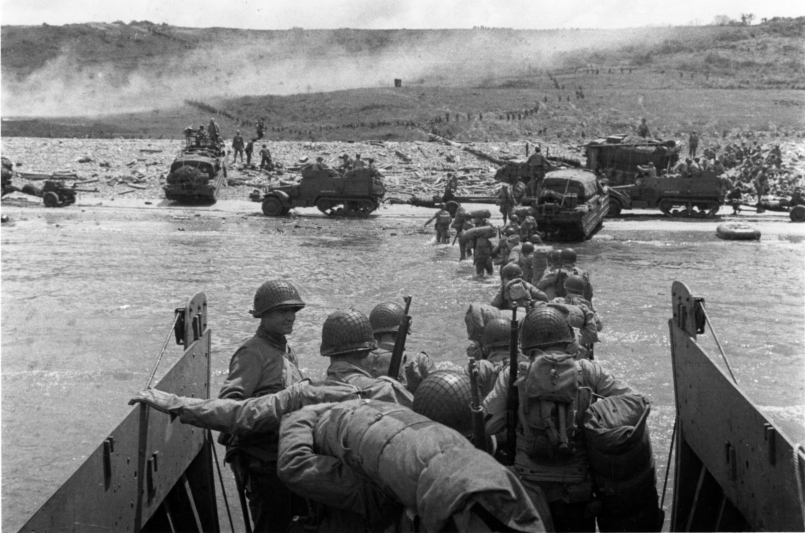 Soldiers in helmets, carrying full packs and arms, are exiting a watercraft and wading in a line towards the beach at Normandy. Not far away, the shoreline is teeming with army vehicles and tanks. Lines of soldiers can faintly be seen cresting a hill on the horizon.