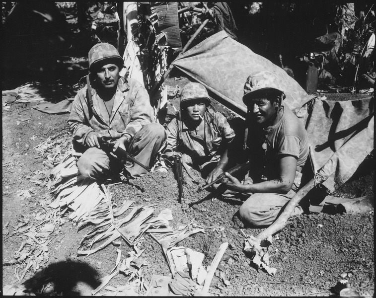 Three Navajo Code Talkers are pictures in army gear. They are sitting on the ground in front of a tent; all holding guns. The man in the middle has a cigarette in his mouth. This is in Saipan, and tropical foliage can be seen in the background.