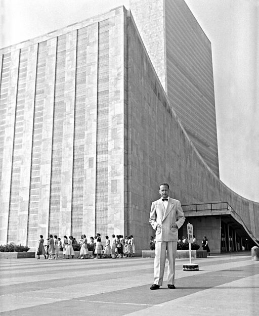 A black and white photo from the 1950s of UN Secretary-General Dag Hammarskjöld standing in front of the General Assembly building wearing a white suit and black bowtie.