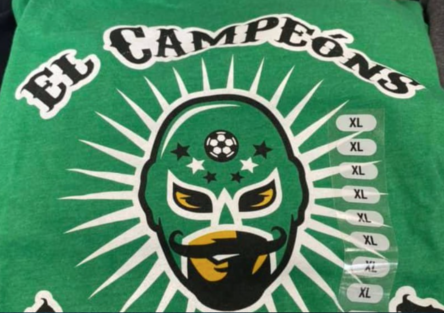 A t-shirt with the phrase "El Campeóns" and an image of a face wearing a luchador mask and a mustache.