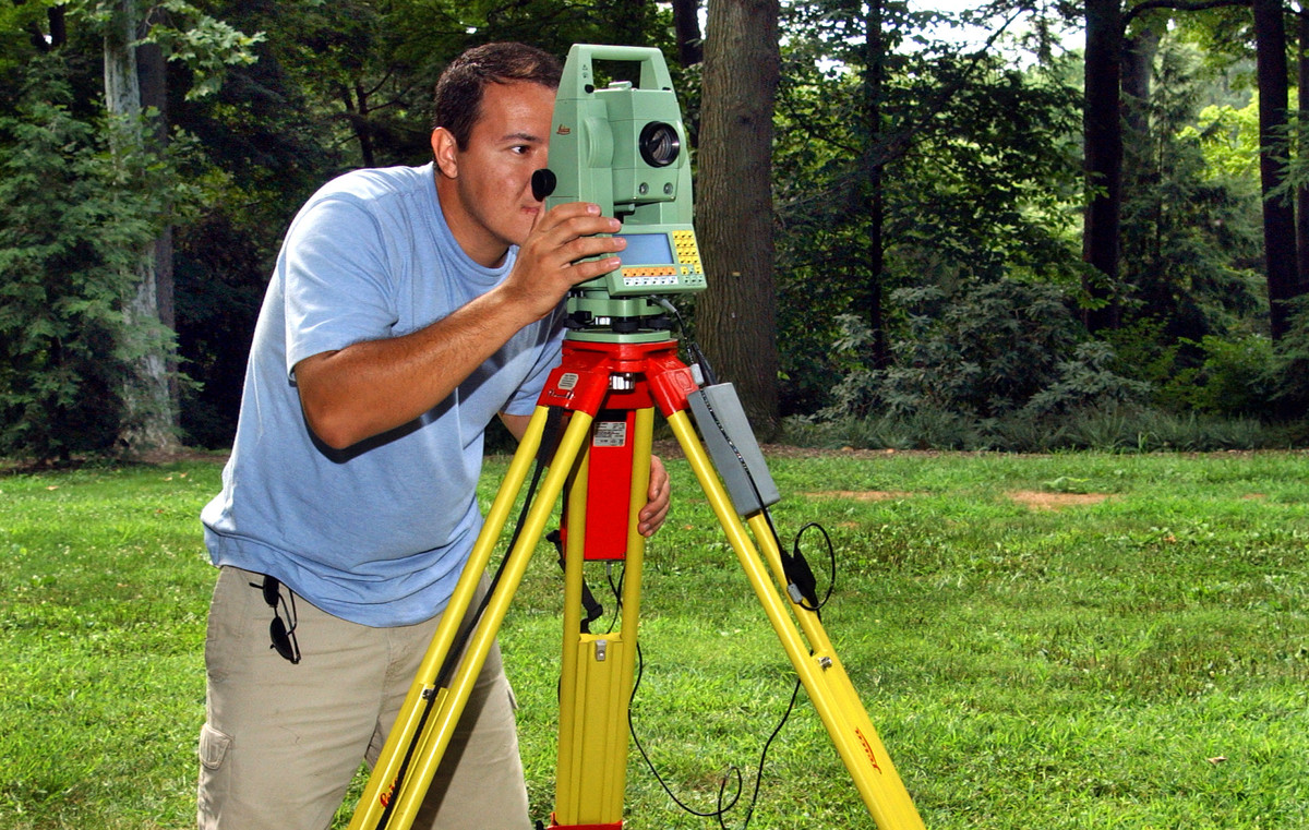 A person at the edge of the woods looks through surveying equipment mounted on a tripod