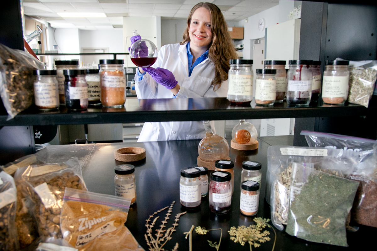A person in a lab coat holds a bottle of dark liquid while standing behind a lab table covered in various plant parts and jars.