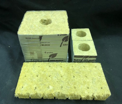 A square, dense block of white mineral wool with a hole in the center in the back left.  Two smaller squares that have holes in the center in the top right.  A large slab of mineral wool lies in front of both blocks.