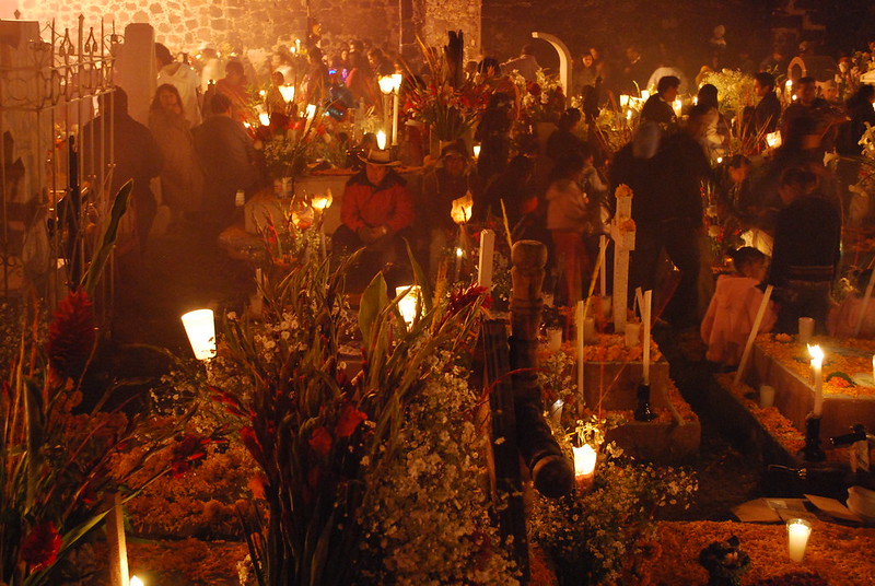 A crowd of people gathered in a cemetary at night.  Graves are decorated with plants and flowers and there are lights throughout.